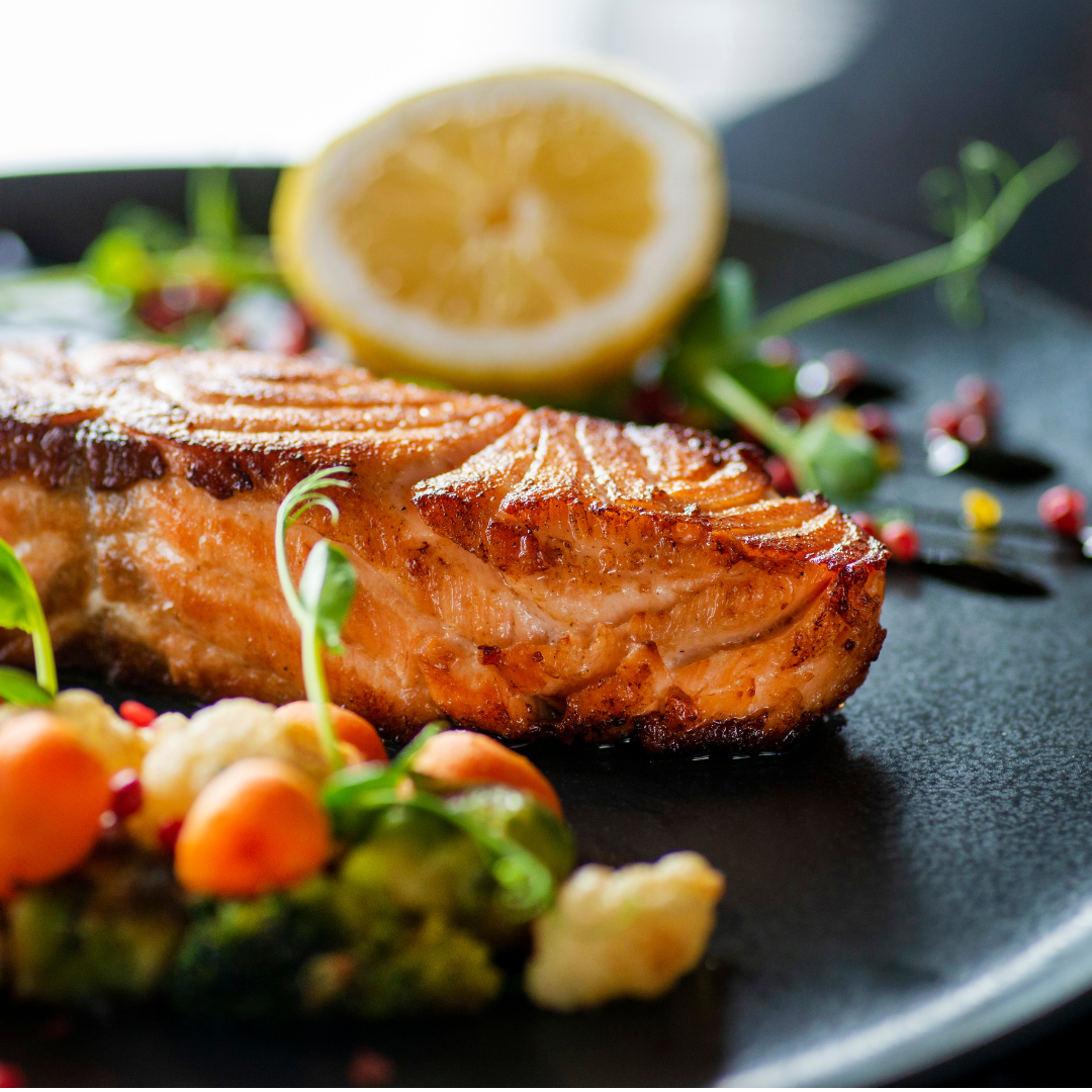 Seared salmon fillet with crispy golden skin served on a dark plate beside steamed broccoli, cauliflower, carrots, a lemon slice, and pink peppercorns.