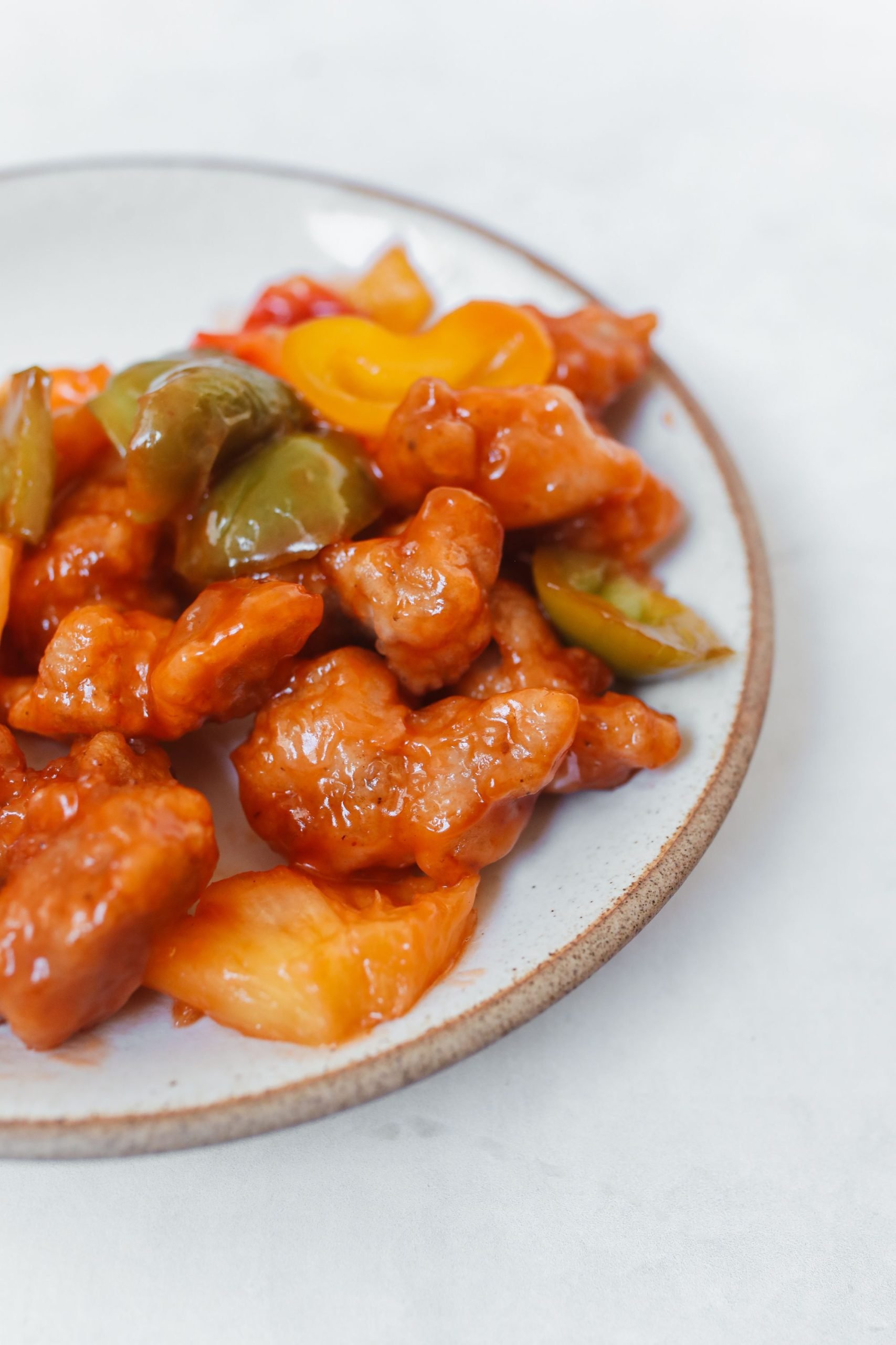 Crispy fried chicken pieces glazed in red sweet and sour sauce with chunks of colorful bell peppers on a white plate.